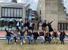 A fun picture of the IDEP student group celebrating in front of Gund Hall, home to the Harvard Graduate School of Design. Nine students are seated in the front row, with eight standing in the back. Most students are smiling, laughing, or throwing their hands up in the air.