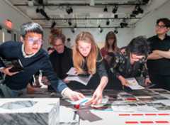 Six people around a table reviewing and sorting printed materials. The focus is on a man and woman reaching across the table to place it in an open spot.