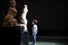 In a dark gallery of ancient sculptures, a young boy looks up at a marble torso in wonderment