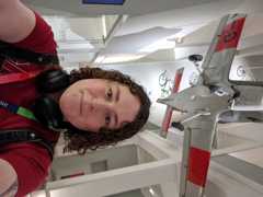 A girl with curly hair (who is quite tall) with a red t-shirt with the Armakuni logo on, headphones around her neck and a pinafore dress on, standing under a plane suspended from the ceiling at Agile Scotland at the National Museum of Scotland