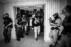 A group of people in a hallway. Some of them are masked and armed.