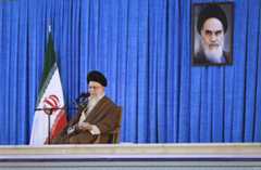 Ali Khamenei, kledd i turban holder en tale. Et bilde av Ruhollah Khomeini henger på veggen bak ham.