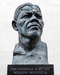 Bildet viser en byste av Nelson Mandela, laget av bronse. Bysten er plassert mot en lys bakgrunn, og den viser et ansikt med tydelige trekk. Han har kort, krøllete hår og et bestemt uttrykk. Under bysten står det en mørk plate med inskripsjonen: "THE STRUGGLE IS MY LIFE" og navnet "NELSON MANDELA."