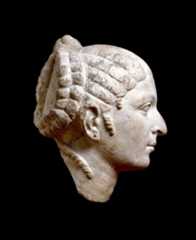 Foto av et skulpturhode i profil, vendt mot høyre. Hodet er utskåret i lys stein og har detaljerte trekk, inkludert en markert nese, små lepper og et rolig uttrykk. Håret er arrangert i tykke, flettelignende seksjoner som er trukket bakover og samlet i en knute. Figuren har også en liten ørepynt. Bakgrunnen er helt svart, noe som fremhever formen og detaljene i skulpturen.