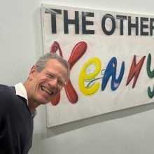 A man with glasses and a big smile posing in front of a colorful painting with the words “THE OTHER KENNY”