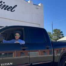 Man wearing sunglasses driving a black Nissan Titan pickup truck parked in front of a white building with the word 'Sentinel' on the wall under a clear blue sky.