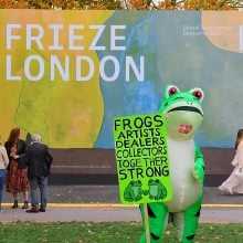 A person in a frog costume holds a bright green sign reading “FROGS ARTISTS DEALERS COLLECTORS TOGETHER STRONG” in front of the entrance sign for Frieze London.