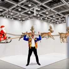 A bright, empty art-fair booth with white partition walls and grid lighting overhead. A cut-out figure stands center with arms raised, while a cartoon Santa in a sleigh and reindeer fly across the background. The wall on the right reads “Art Basel.”