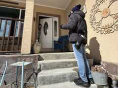 A person in winter clothing waits on the steps outside a house as someone else peeks their head out of the door