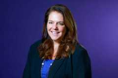 A woman with shoulder-length brown hair smiles against a purple backdrop. She wears a black blazer over a royal blue top and a silver necklace.