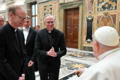 Two priests in black suits and clerical collars smile at Pope Francis within an ornate Vatican hall.