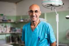 A bald man with glasses and a beaded necklace, wearing blue medical scrubs, smiles in a clinic. The light green room has shelves of supplies, a surgical lamp, and a wooden crucifix on the wall.