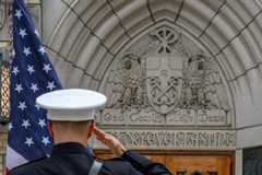 A person in a dark uniform and white hat salutes the American flag outside the Basilica of the Sacred Heart. Stone carvings above the door feature eagles, a shield, and "God Country Notre Dame."