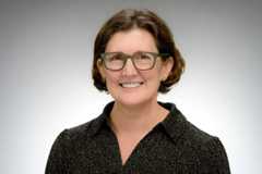 A woman with short brown hair, olive green glasses, and a black collared shirt with light brown dots smiles at the camera.