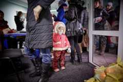 A small Ukrainian child in a red winter coat with cream fur, red pants with gold butterflies, and light blue gloves stands looking forward, sucking a gloved thumb. Adults in winter clothes fill a crowded indoor waiting area, with others reflected in a glass door. Bags are on the floor.