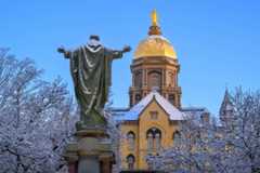 Snow-dusted Sacred Heart of Jesus statue with arms outstretched, seen from the back. Behind it, Notre Dame's Golden Dome Main Building and snow-covered trees are under a bright blue sky. (Main Quad on a snowy morning)