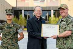 A U.S. Marine in uniform stands at attention next to a priest in a black coat and collar, who is smiling while accepting a framed Department of Defense certificate from a smiling U.S. Navy service member in camouflage uniform. They are outdoors in a courtyard with campus buildings in the background.