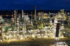 An aerial view of a vast industrial refinery, brightly lit at night. Thousands of yellow lights illuminate the intricate network of towering columns, pipes, and buildings under a dark blue sky, with visible steam plumes.
