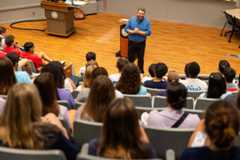 A bearded man in a blue checkered shirt smiles with clasped hands at a lectern, addressing students in tiered seating within a Notre Dame lecture hall.