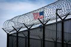 Razor and barbed wire atop a fence near the Saint Joseph County Jail in South Bend, with American flag flying in the background.