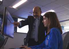 Photo: Professor points at a computer screen while a seated student looks on.
