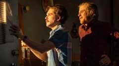 Two men converse, one holding a bottle, in a dimly lit room.