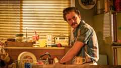 A man with a moustache stands in a kitchen, wearing a blue striped shirt.