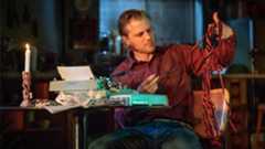 A man examines tangled wires at a table, with a candle illuminating the scene.