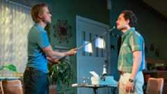 Two men confront each other, one holding a stick, in a green-walled room.