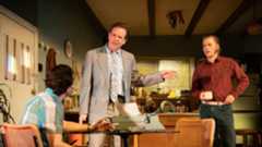 Three men interact in a warmly lit room with retro decor and furniture.