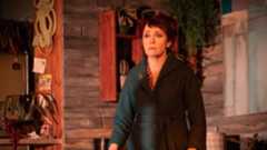 A woman in a dark green coat stands in a rustic, dimly lit room.