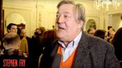 A man in a grey coat speaks, surrounded by a crowd in a theatre.