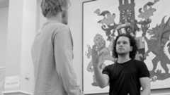 Two men stand facing each other, with a colourful mural in the background.