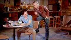 Two men converse at a table in a cluttered kitchen setting.