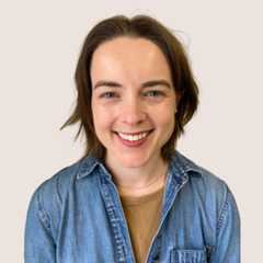 A person with short brown hair is smiling at the camera. They are wearing a denim shirt over a light brown top, and the background is plain and light-colored.