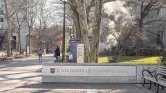 A stone sign reading University of Pennsylvania stands at the entrance to a campus walkway lined with trees, benches, and street lamps, with people walking and smoke rising in the background.