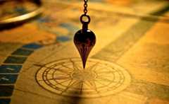 An image of a flat, golden compass, with a pointed pendulum hanging over it.