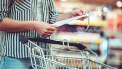 Eine Person in einem gestreiften Hemd steht in einem Geschäft neben einem Einkaufswagen und prüft einen langen Kassenbon. Die Regale mit den Produkten sind im Hintergrund unscharf.