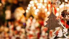 Im Vordergrund hängt hölzerner Christbaumschmuck, im Hintergrund leuchten warme, verschwommene Weihnachtslichter und festliche Dekorationen.