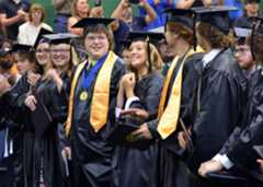 A female graduate grips her diploma and smiles as male and female graduates smile, clap, and look at each other from the front row during the graduation ceremony after turning their tassels.