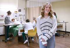 A young female college students smiles, as a Nursing instructor and students complete a class exercise by a medical manikin in a hospital bed.