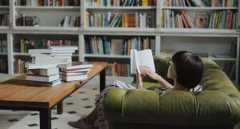 a photo of a woman reading on a couch with bookshelves in the background and stacks on books on a table