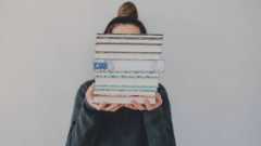 a fair-skinned femme-looking person holding a stack of books that is obscuring their face