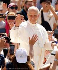 Pope Leo XIV stands and waves, surrounded by a crowd of people holding phones