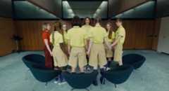 A still of the film Club Zero showing a group of students in uniform inside a classroom space joining hands in a circle with Wasikowska’s character.