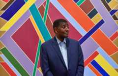 An older Black man wearing a navy suit jacket over a blue checkered button up shirt looks to the side standing in front of a multicolored mural