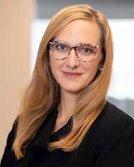 Christina Stephenson has long blonde hair and is wearing glassess and a black suit.