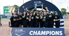 Members of the Talons, wearing their black t-shirts with "Champions" across the chest in green, yellow, and white lettering, and green hats, cheer as they raise the AUSL Championship trophy at Rhoads Stadium behind a Champions sign.