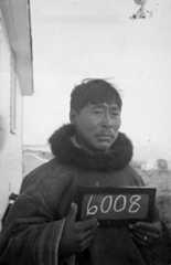 A black-and-white photograph of an Inuit man holding a small chalkboard with the number 6008.