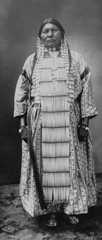 A black-and-white photo of a Dakota First Nation woman wearing a striped dress under a long beaded breastplate. It is matched with a necklace, long earrings and braids.